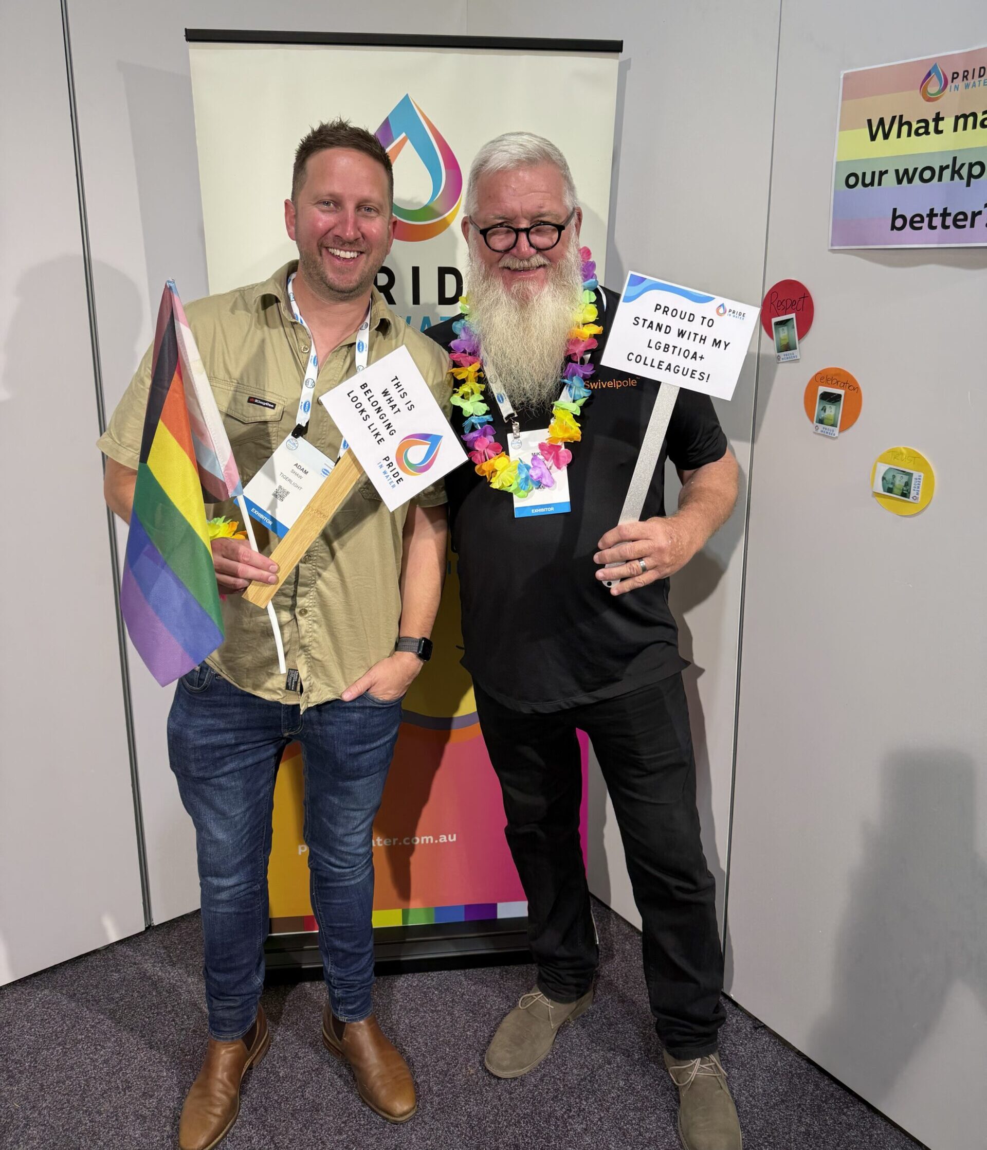 Two smiling conference attendees standing in front of the Pride in Water LGBTIQA+ Network pull-up banner at an industry exhibition. One holds a Progress Pride flag and a sign reading "This is what belonging looks like." The other wears a rainbow flower lei and holds a sign reading "Proud to stand with my LGBTIQA+ colleagues!" The interactive rainbow wall activity is visible behind them.